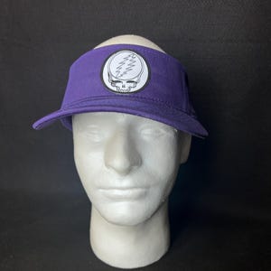 May include: A purple visor with a white and black emblem featuring a skull and lightning bolt design. The visor is displayed on a white mannequin head against a black background. The visor has a curved brim.