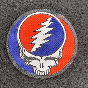 Puede incluir: Un parche redondo con un diseño de calavera estilizada y un rayo. El diseño está dividido en secciones rojas y azules, con una calavera blanca y un rayo en el centro. El parche tiene un borde negro.