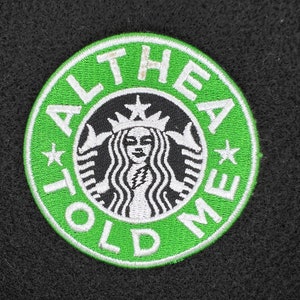 Puede incluir: Un parche bordado circular verde y blanco con el texto "ALTHEA TOLD ME" alrededor de un logotipo en blanco y negro. El logotipo presenta el rostro de una mujer y una corona.
