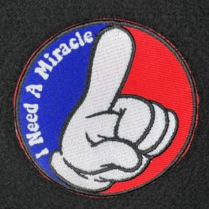 May include: A round, blue and red embroidered patch with a white hand giving a thumbs up. The text "I Need A Miracle" is written in white on the blue portion of the patch.