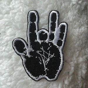 Peut inclure: Patch brodé noir représentant une main faisant le geste rock and roll avec un motif d'étoile blanche à l'intérieur de la paume.