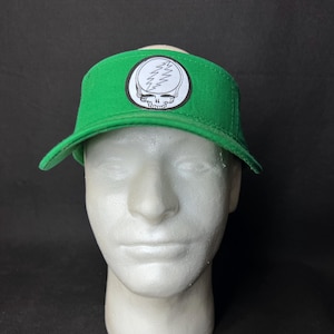 May include: Green visor with a white and black skull and lightning bolt emblem. The visor is made of a durable material and has a curved brim. The emblem is circular and prominently displayed on the front of the visor.
