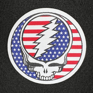 Puede incluir: Un parche redondo con un contorno blanco y una imagen en blanco y negro de una calavera con un rayo en su interior. La calavera está superpuesta sobre una bandera estadounidense roja, blanca y azul.