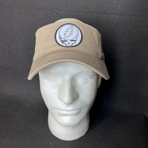 May include: Tan visor featuring a white and black Steal Your Face logo. The logo is a skull with a lightning bolt through it, set inside a white circle. The visor is displayed on a white mannequin head.