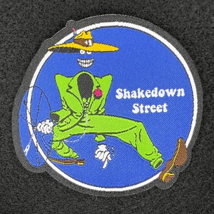 May include: An embroidered patch featuring a cartoon character in a green suit and hat, holding a whip. The text "Shakedown Street" is embroidered in white on a blue background.