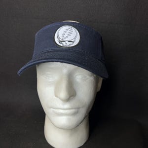 May include: Navy blue visor cap with a white and black patch featuring a skull and lightning bolt design. The cap has a curved brim and is displayed on a white mannequin head.