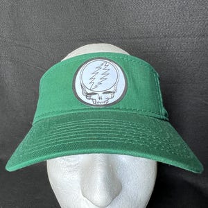 May include: Green visor featuring a white and black graphic of a skull with lightning bolts. The visor has a curved brim and is made of a durable fabric. The graphic is centered on the front of the visor.