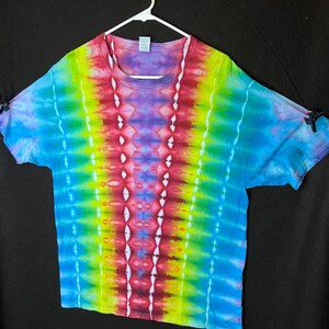 May include: A colourful tie-dye t-shirt in a rainbow of shades, including red, yellow, green, blue, and purple. The shirt is displayed on a white hanger against a black backdrop. The tie-dye pattern is vertical with a white design down the centre.
