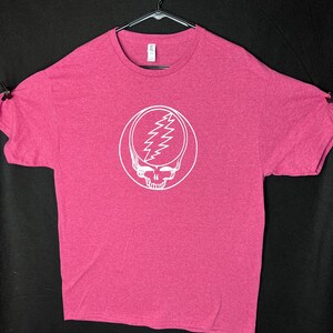 May include: A cerise t-shirt featuring a white graphic of a skull with a lightning bolt design. The shirt is displayed on a black hanger against a black backdrop. The tee has short sleeves and a round neck.