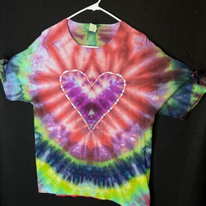 May include: A tie-dye t-shirt with a large, white-outlined heart in the centre. The heart is purple and pink, and the shirt is a mix of red, pink, green, blue, and yellow.