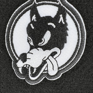 Op de afbeelding: Geborduurde patch met een cartoon wolf ontwerp in zwart en wit. De wolf heeft een brede grijns en steekt zijn tong uit. De patch is geplaatst in een witte cirkel met een zwarte rand, op een zwarte achtergrond.