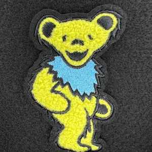 Op de afbeelding: Geborduurde patch met een gele beer met een blauwe kraag en zwarte omtrek. De beer heeft een lachend gezicht en staat rechtop. De patch staat op een zwarte achtergrond.