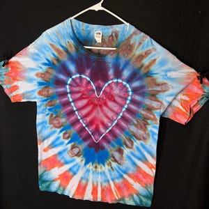 May include: A tie-dye t-shirt with a white heart design in the centre. The shirt is a mix of blue, red, orange, and brown colours.