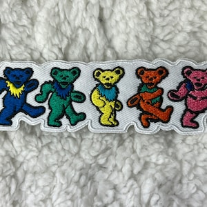 Peut inclure: Écusson brodé représentant cinq ours dansants de couleurs vives : bleu, vert, jaune, orange et rose. Les ours sont bordés de blanc et se détachent sur un fond blanc.