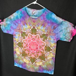 May include: A colourful tie-dye t-shirt with a central starburst design. The shirt's pattern features pink, green, and white, surrounded by blue, purple, and yellow. The short-sleeved shirt is displayed on a white hanger against a black background.