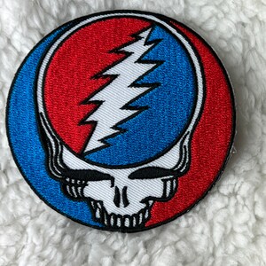 May include: A circular embroidered patch with the Steal Your Face skull logo. The design features a red and blue split background, a white lightning bolt, and a white skull.