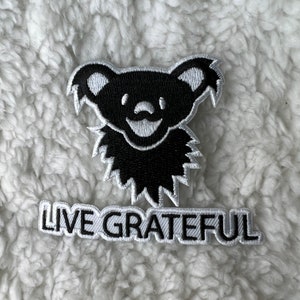 Peut inclure: Écusson brodé noir et blanc représentant un ours stylisé souriant. En dessous, une inscription rectangulaire blanche avec les mots "LIVE GRATEFUL".