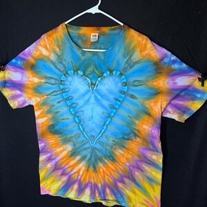 May include: A tie-dye t-shirt with a blue heart design in the centre. The shirt is a blend of orange, yellow, purple, and blue colours.