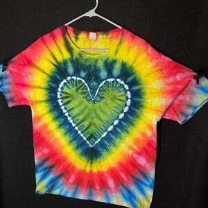 May include: A tie-dye t-shirt with a white heart design in the centre. The shirt is a blend of red, yellow, blue, and green colours.
