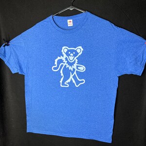 May include: A blue t-shirt featuring a white dancing bear design. The short-sleeved top is suspended on a black hanger against a dark background. The bear graphic has a smiling face and is outlined in white.