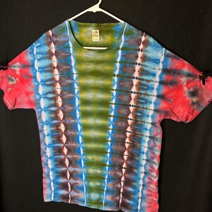 May include: A tie-dye t-shirt with a crew neck, displayed on a white hanger. The shirt features a vibrant pattern of blue, green, red, and brown vertical stripes. The sleeves are short and the shirt is made of a soft material.