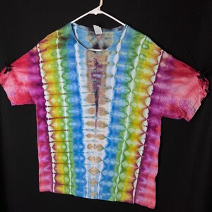 May include: A vibrant tie-dye t-shirt in a rainbow pattern, displayed on a white hanger against a black backdrop. The shirt showcases vertical stripes of pink, red, yellow, green, blue, and purple. The sleeves are finished with black ties.