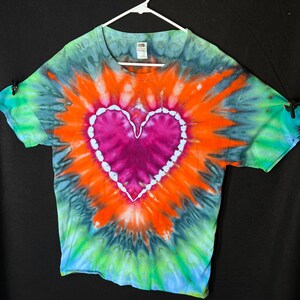 May include: A tie-dye t-shirt featuring a large pink heart at the centre. The heart is outlined in white and the shirt is a blend of blue, green, orange, and black.
