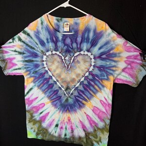 May include: A tie-dye t-shirt with a white heart design in the centre. The shirt is a blend of purple, blue, green, pink, and yellow colours.