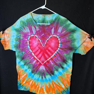 May include: A tie-dye t-shirt featuring a large, pink heart design in the centre. The shirt is a blend of blue, green, orange, and purple colours.
