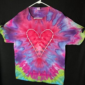 May include: A tie-dye t-shirt with a white heart design in the centre. The shirt is a mix of pink, purple, blue, green, and yellow.