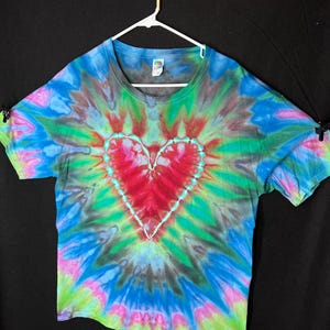 May include: A tie-dye t-shirt with a large red heart in the centre. The shirt is a blend of blue, green, and pink colours.