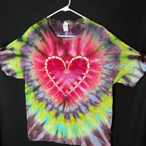May include: A tie-dye t-shirt with a large heart design in the centre. The shirt is a blend of pink, red, green, blue, and black colours.