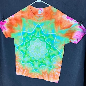 May include: A tie-dye t-shirt with a vibrant starburst design. The shirt features a blend of orange, green, blue, and pink hues. The short-sleeved tee is hanging on a white hanger against a dark background.