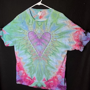 May include: A tie-dye t-shirt with a large purple heart in the centre. The shirt is a mix of green, blue, pink, and purple colours.