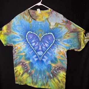 May include: A tie-dye t-shirt with a large, white-outlined heart in the centre. The shirt features a vibrant blend of blue, yellow, and purple hues, creating a colourful, abstract design. The shirt is hanging on a white hanger against a black background.