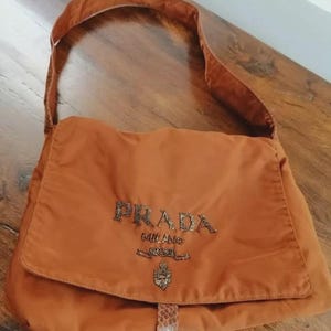 May include: A rust-colored shoulder bag featuring a flap closure with a single strap. The front displays the text PRADA MILANO and a crest logo embellished with small dark beads. A textured, snakeskin-patterned tab secures the front flap closure.