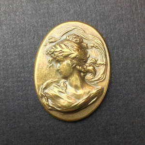 May include: A gold-toned oval-shaped metal charm featuring a raised relief design of a woman's profile wearing a wreath and flowing drapery.