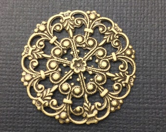 May include: A gold-toned metal filigree embellishment with a circular design. The embellishment features a central flower with intricate scrollwork and small round accents.