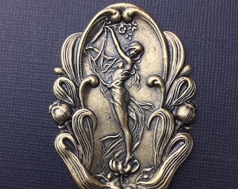 May include: A gold-toned metal ornament featuring a stylized female figure holding a flower. The figure is surrounded by ornate floral details and is set against a dark background.