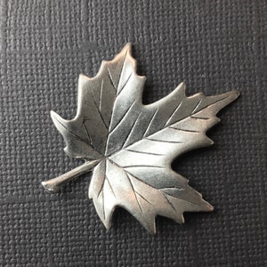 Könnte beinhalten: Eine silberfarbene Ahornblatt-Brosche mit einem detaillierten, strukturierten Design.