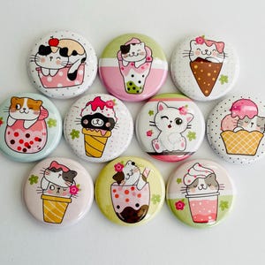 May include: A collection of ten round, colorful pinback buttons featuring cartoon cats with desserts. The cats are depicted with ice cream, boba tea, and in waffle cones. The buttons have a white background with a variety of colors.