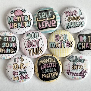 May include: Ten round button pins with positive affirmations. The buttons are white with black, pink, blue, and yellow accents. The text on the buttons includes: "Mental Health", "Self Love", "Take a Deep Breath", "Be Kind to Your Mind", "You Got This", "You Matter", "Be The Change", "It's O.K. to not be O.K.", "Mental Health Does Matter", and "You Are Enough".