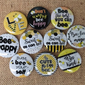 May include: Ten round, yellow and black buttons with bee-themed sayings. The sayings include "Bee Happy", "Sweet as can bee", "Bee-lieve in magic", "Don't worry bee happy", "Bee the best you can bee", "Just bee yourself", "As cute as can bee", "Bee kind", and "Love".