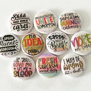May include: Ten 1-inch button pins with various sarcastic sayings. The buttons are white with colorful text and designs. Some of the sayings include "Shhh... No One Cares", "Procrastinators Unite! Tomorrow", "If Only Sarcasm Burned Calories", "Speak Fluent Sarcasm", "No Idea What I'm Doing", "Sassy Since Birth", "Nope Not Today", "If You Love Me Let Me Sleep", "Fresh Sarcasm Served Daily", and "I May Be Wrong But I Doubt It".