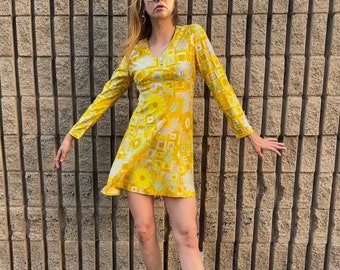 May include: A vintage-style yellow mini dress with a floral and geometric pattern. The dress has long sleeves and a V-neck. The person is wearing white platform heels and sunglasses. The background is a textured, vertical brick wall.