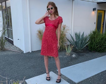 May include: A woman wearing a red lace dress with a red satin sash and black heeled sandals. She is standing in front of a white building with a wooden pergola.