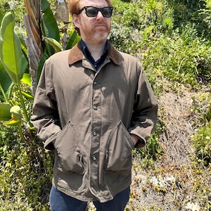May include: A brown jacket with a brown corduroy collar, a button-front closure, and two large front pockets. The jacket is worn over a dark blue collared shirt, paired with dark blue trousers and black sunglasses.