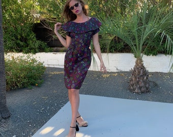 May include: A woman wearing a dark purple dress with a floral print and a ruffled neckline. She is wearing sunglasses and standing on a white surface.