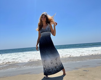 Puede incluir: Una mujer con un vestido largo de maxi azul y blanco con estampado floral se encuentra en una playa de arena con el océano de fondo.