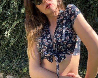 May include: A woman wearing a dark blue floral print crop top with a tie front and a light blue skirt. She is wearing green sunglasses.